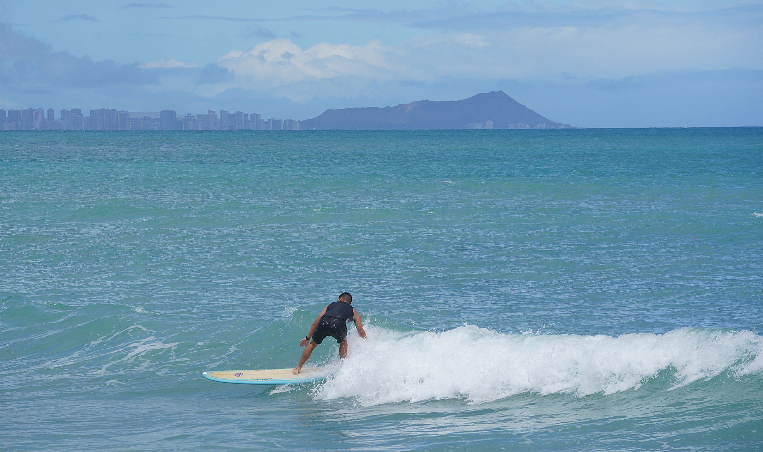 Man surfing in Ewa Beach, with the Honolulu skyline and Diamond Head far off in the distance. 