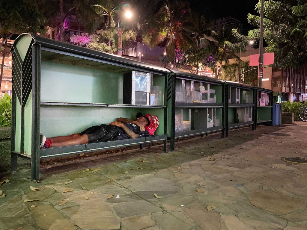 Person sleeps in magazine racks along Kalakaua Avenue in Waikiki.