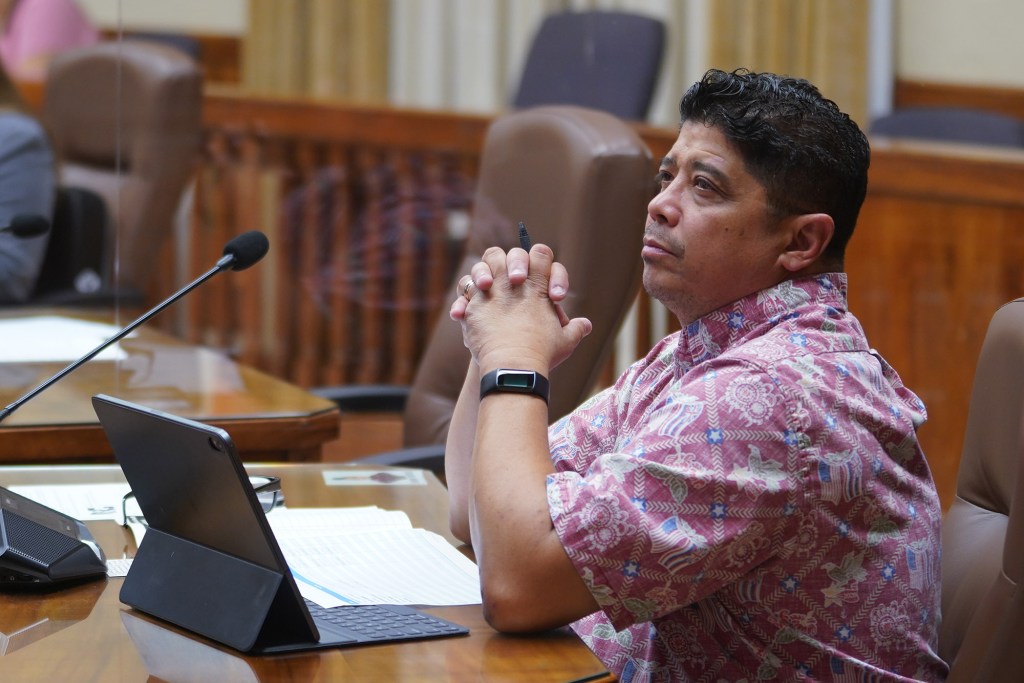Honolulu City Council member Augie Tulba listens to public testimony on a large video screen at Honolulu Hale.