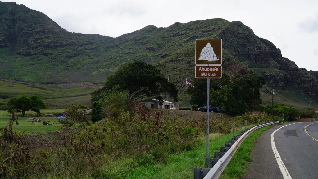 A road sign indicating Ahupua‘a Makua is in the foreground, with soldiers and an American flag in the distance.