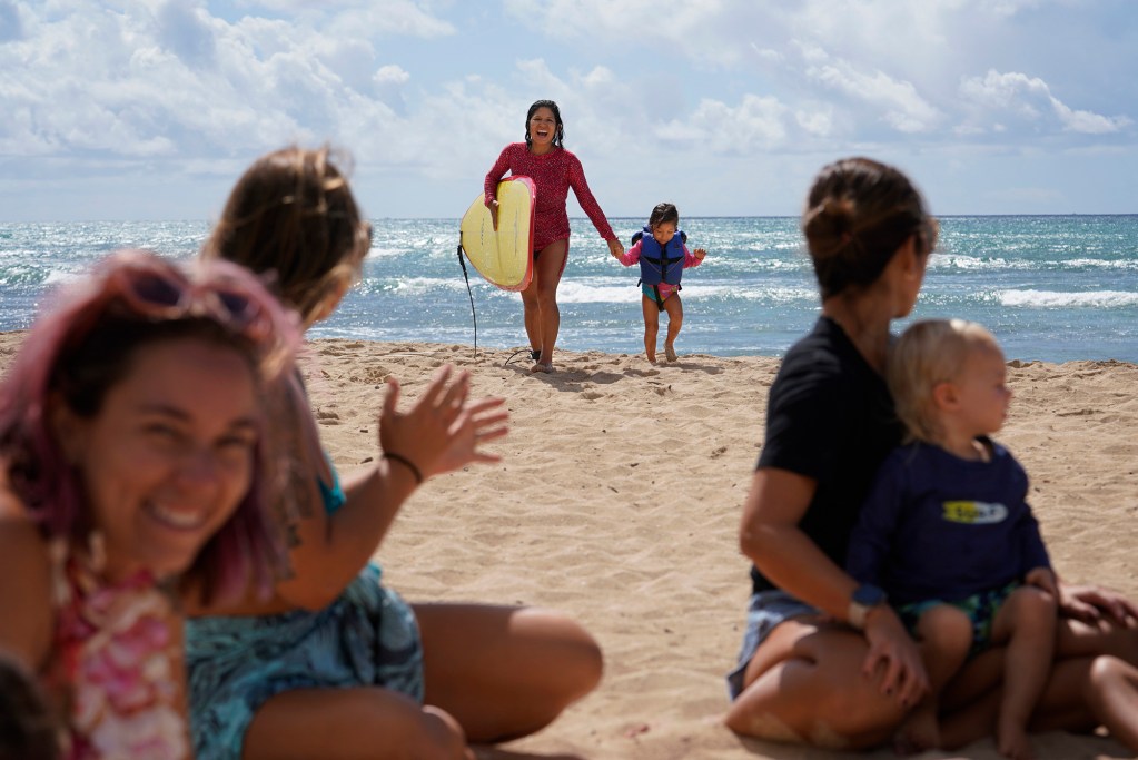 Surfing Mom’s Melissa Martin and her daughter Esperanza return after surfing while Erica Diehl, from left, Victoria Moody, Sachiko Stokes and Daniel Ritchie play on the sand Tuesday, Aug. 15, 2023, at White Plains Beach in Ewa Beach. The moms take turn surfing while other moms watch the keiki. (Kevin Fujii/Civil Beat/2023)