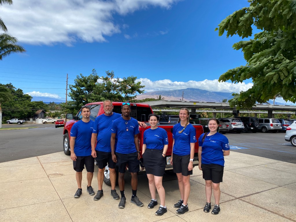 Six people in matching blue shirts stand in front of a pickup truck, with the mountains of West Maui looming in the background 
