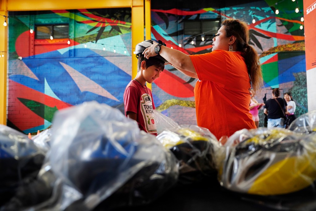 Keegan Cooper, left, 10 of Kaneohe, tries on a helmet with the help of volunteer Jacqueline Dardeen of Ewa Beach as part of Hawaii Bicycle League Bike Month 2024, HBL hosts a helmet giveaway Sunday, July 28, 2024, at SALT at Kakaako in Honolulu. 300 helmets were available to give away to adults and keiki. Hawaii state law requires those younger than 16-years-old to wear a helmet while riding a bicycle or scooter. (Kevin Fujii/Civil Beat/2024)