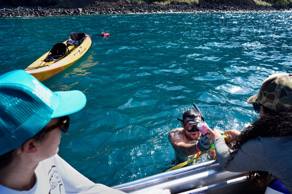 Jetaimoio Henshaw, left, watches boatman Kahina Hewitt had her partner Swope Conn drinking water for his team during the Kealakekua Bay Non-Native I‘a (fish) Removal Event Sunday, May 18, 2025, in Captain Cook. Henshaw said, “You’re in the biggest world’s biggest desert so drink up!” (Kevin Fujii/Civil Beat/2025)