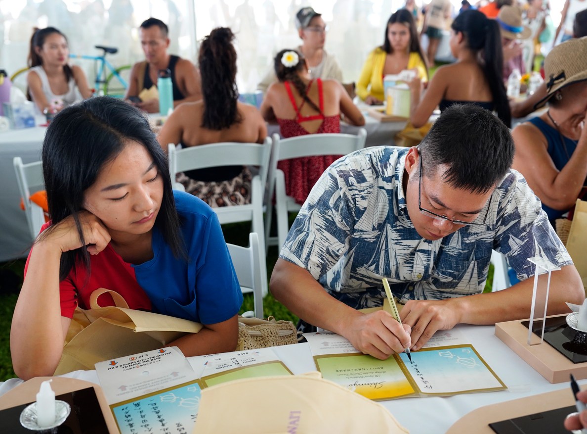 Honolulu residents Qiyu Yin, left, watches Shintzu Jen write in traditional Chinese for his 27th Annual Shinnyo Lantern Floating Hawaiʻi at Ala Moana Regional Park Monday, May 26, 2025, in Honolulu. 6,000 lanterns with messages to lost loved ones are launched by family members. (Kevin Fujii/Civil Beat/2025)