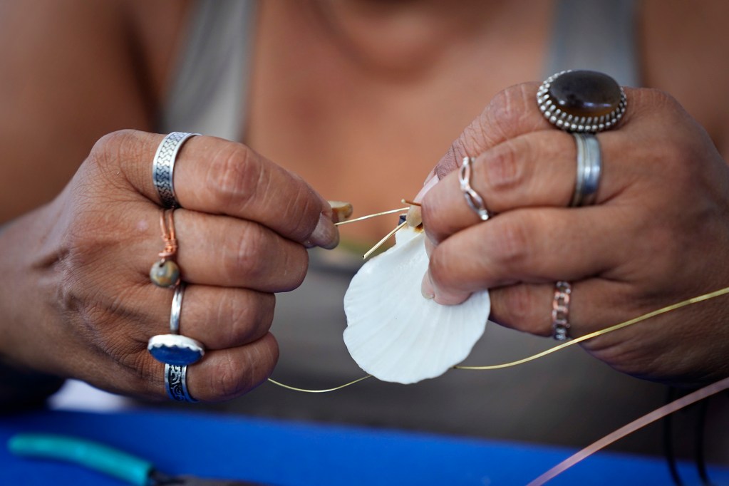 808 N8iv jewelry maker Maile Kalahiki makes a necklace with a seashell while selling her and a friend’s accessories at the Hilo Farmer’s Market Saturday, May 31, 2025, in Hilo. The bi-weekly outdoor market hosts up to 200 vendors selling artisanal crafts, foods and produce. (Kevin Fujii/Civil Beat/2025)
