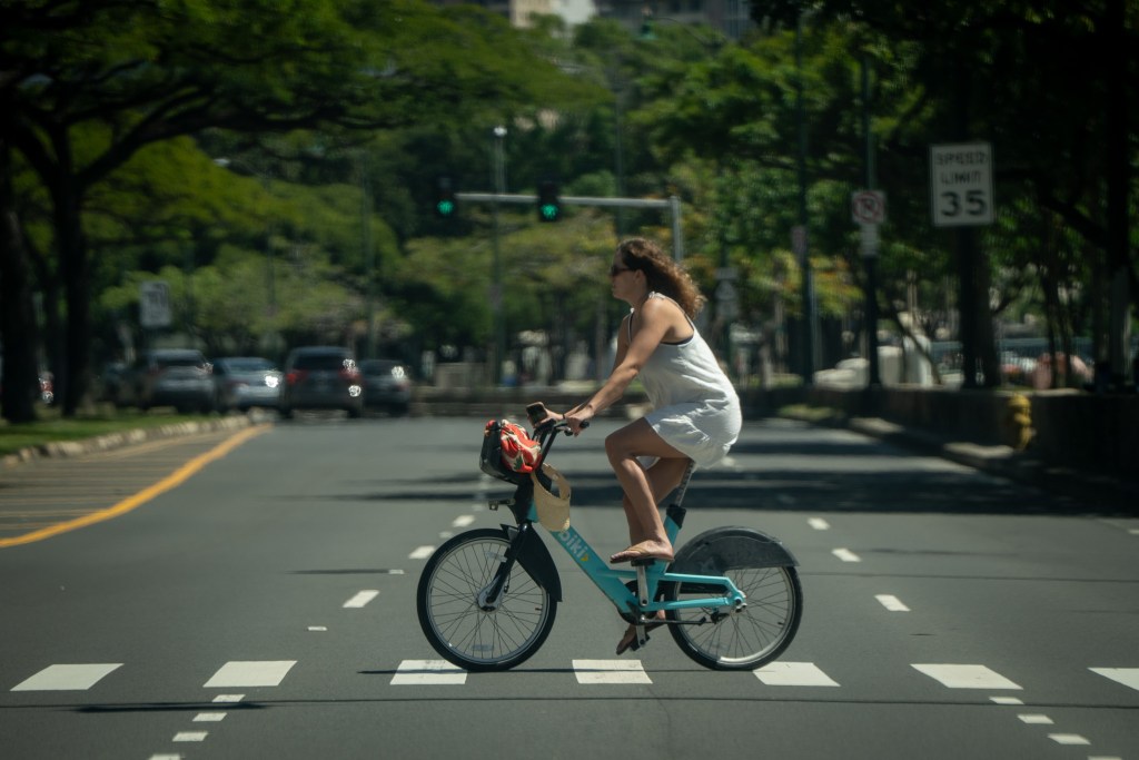 A woman wearing a white outfit bicycles a blue Biki rental bike on a crosswalk. The roadway and foliage recede into the background.