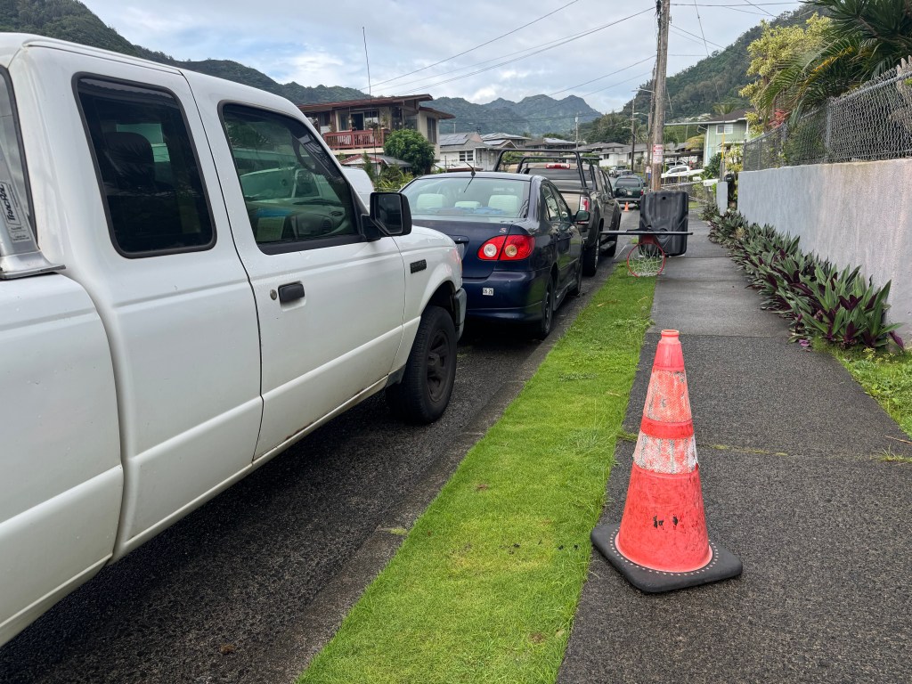 An orange cone sits on the sidewalk next to a car and a truck parked tightly in a Kalihi Valley neighborhood