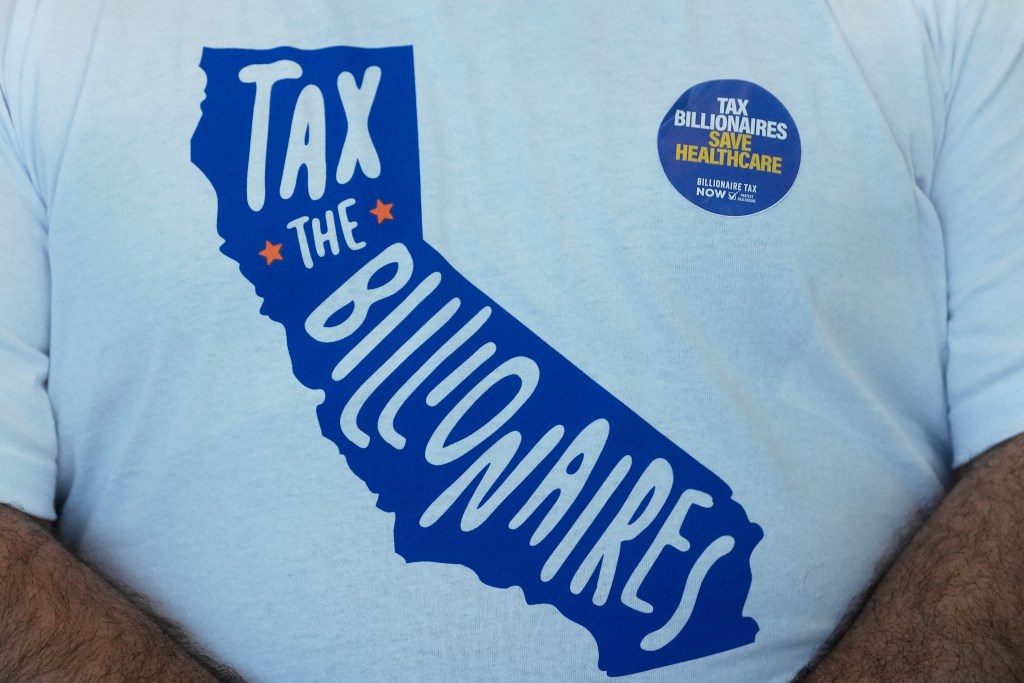 A man's shirt and sticker are displayed at the Billionaire Tax Now booth at the 2026 California Democratic Party State Convention in San Francisco, Saturday, Feb. 21, 2026. (AP Photo/Jeff Chiu)