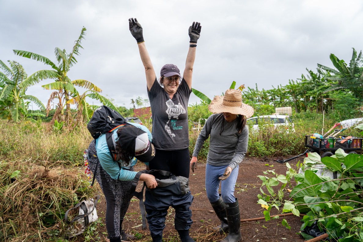 Megan Dooley gets a hand putting on waders during work clearing a ditch of overgrown trees and foliage at Mill 6 Farms in Waialua May 23, 2026. Rebecca Stringer, left,  and Julie Taniyama lend a hand. (Craig Fujii/Civil Beat/2026)