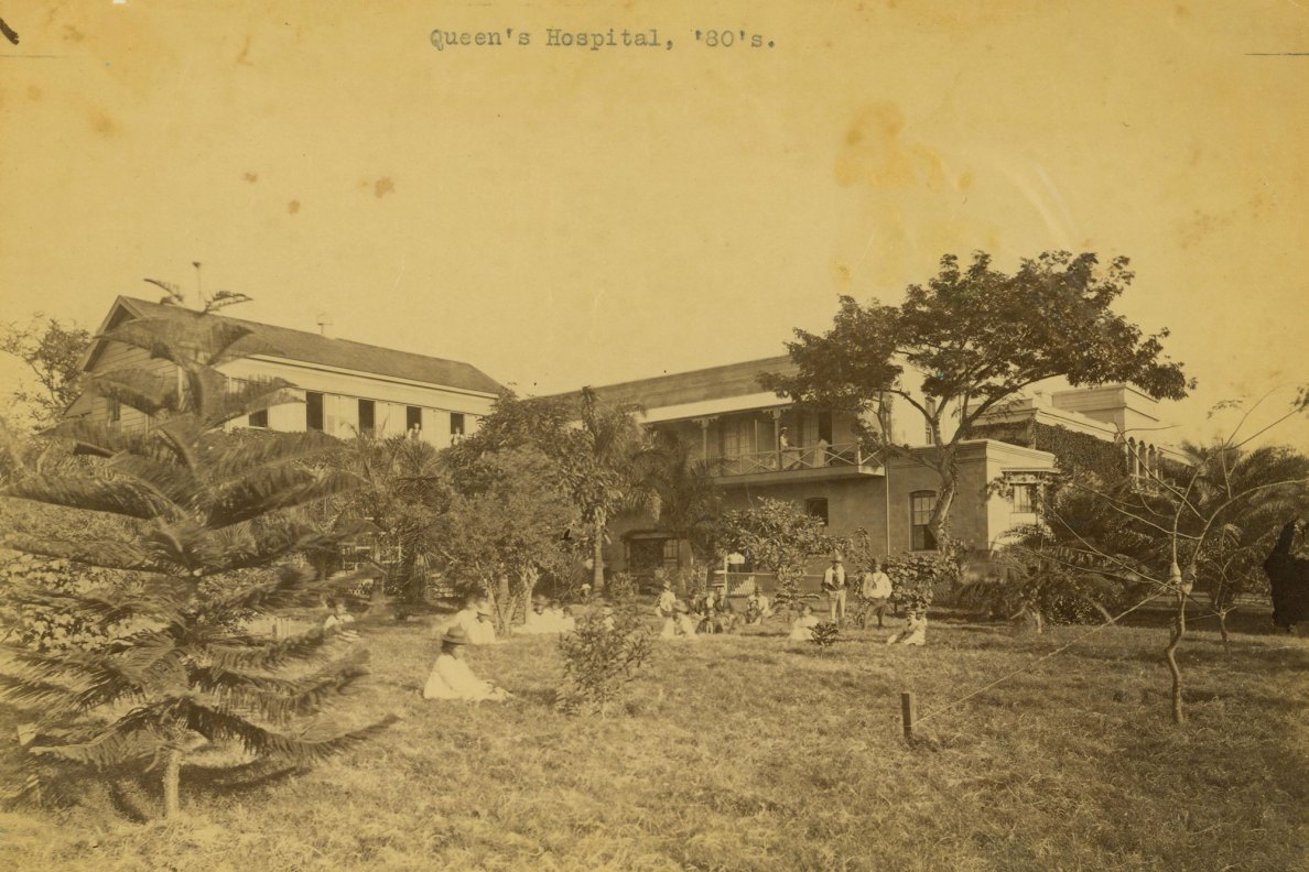 Photo of Queen's Hospital with people sitting on the grassy lawn.