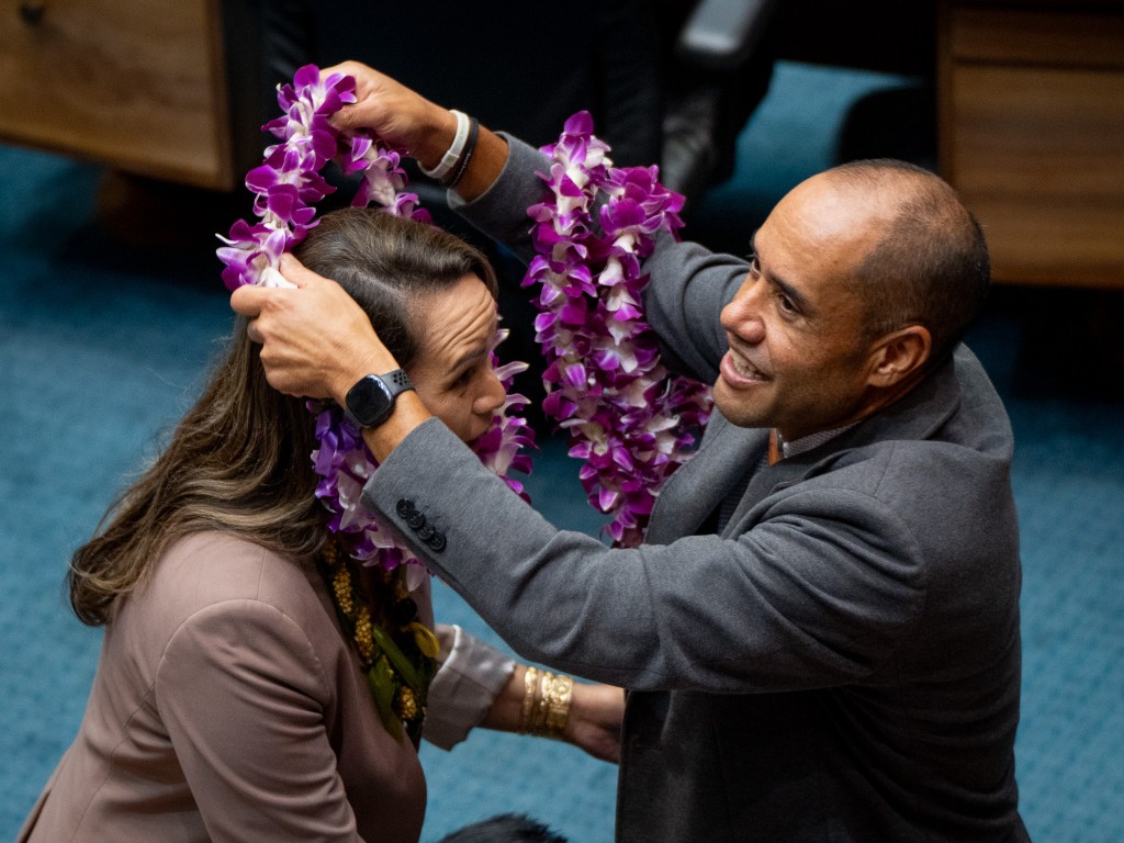 A balding man in a gray blazer, right, places lei on a female with long brown hair and a medium colored blazer.