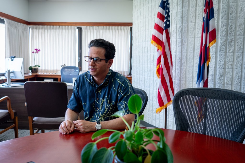 A clean-shaven man wearing glasses and a blue patterned shirt sitting at a table in front of flags and a white brick wall.