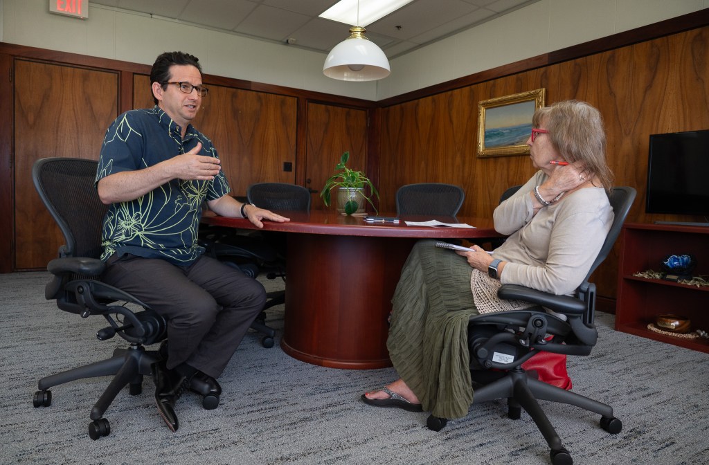 A dark-haired man with glasses and a blue patterned shirt, left, speaks with a woman wearing a beige top and an olive floor-length skirt as they sit at a table in an office.